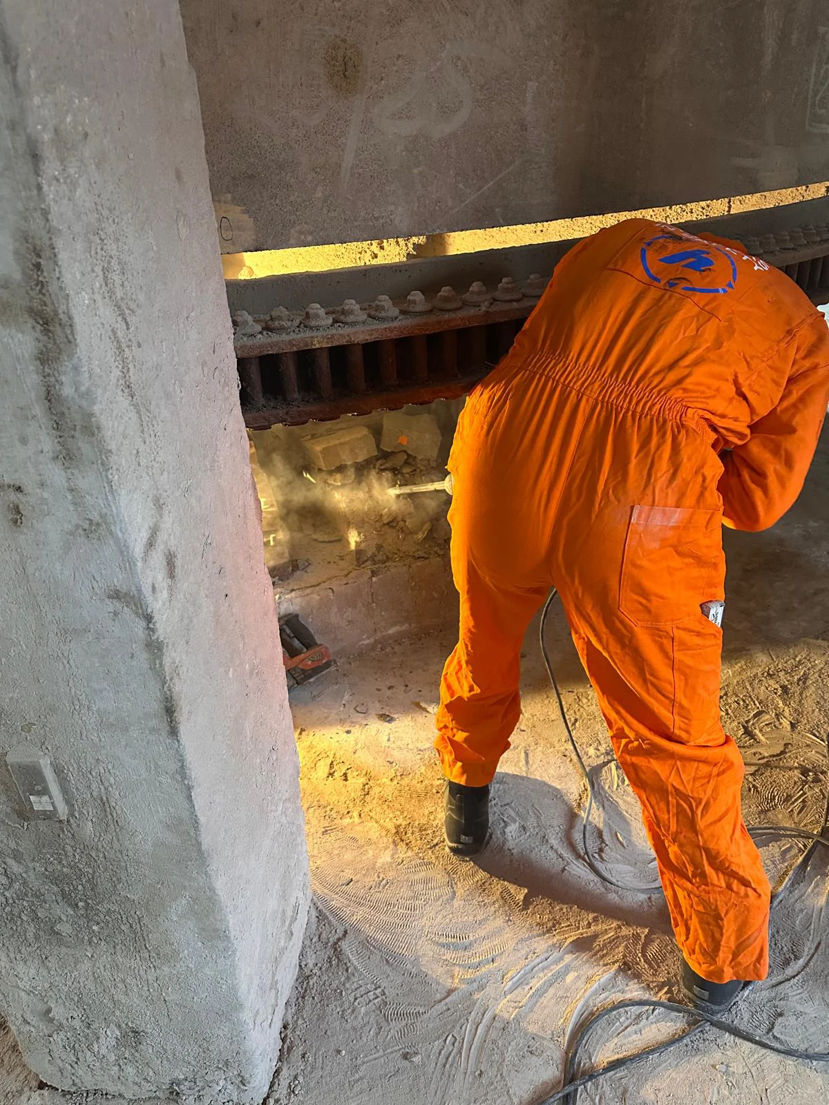 Pracownik budowlany w kombinezonie ochronnym skuwający stary fundament pod wielkim wieńcem zębatym maszyny przemysłowej.